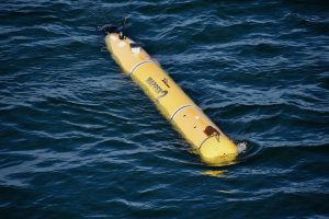 A torpedo-shaped robot with a yellow body and black tail cone bobs at the ocean’s surface. The robot has a label that reads MAPPER 2 in thick black letters. The background is dark-greenish-blue ocean.