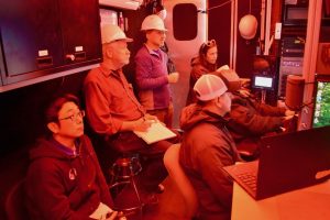 Six researchers watch video monitors during deployment of a robotic submersible. Five of the researchers are seated and one is standing. The researchers are assembled in a control room inside a shipping container. The right-hand side of the control room has video monitors and electronics. The photo has a red tint from the red lights in the control room.