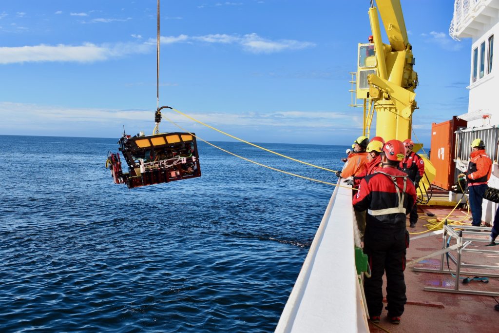 Marine operations crew members launch a robotic submersible into the water. The crew members are aboard a research ship on the right-hand side of the frame. The submersible has a yellow float, black frame, and a red mechanical arm, and is equipped with various scientific instruments. A black tether extending offscreen at the top of the frame is connected to a yellow crane on the research ship to lift the submersible from the ship into the water. Marine operations crew members are holding two yellow tethers, one to carry video from the submersible’s cameras and one to stabilize the submersible during deployment. The background is blue ocean and bright-blue sky with scattered streaks of white clouds.