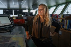 A researcher with long blonde hair wearing a brown scarf, black fleece jacket, and dark-blue jeans stands on the bridge of a research ship. The researcher has her right hand on a counter and her left hand on her hip. A counter with maps, charts, and a navigation display is in the foreground. Shelves with binders, scientific instruments, and video monitors are in the background in front of windows looking out to sea.