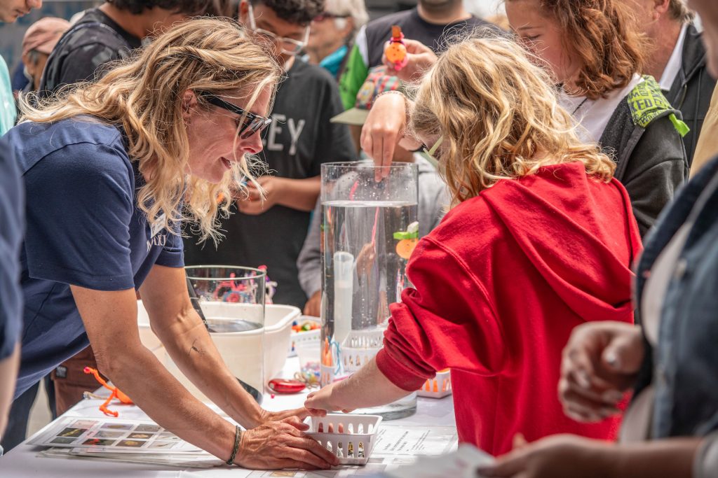 An MBARI researcher with shoulder-length blonde hair, black sunglasses, and a blue t-shirt holds a plastic box of supplies for a young visitor with shoulder-length blonde hair wearing a red hooded sweatshirt. The background is a science demonstration with two transparent plastic cylinders of water and a crowd of people.