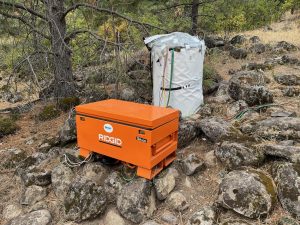 MBARI’s cylindrical 2G ESP instrument, covered in a white casing, installed in the forest. The 2G ESP instrument is positioned between large dark-brown boulders covered in dead pine needles and greenish-brown moss. A series of tubes and wires extend out of the frame to the right. In the foreground is a large, rectangular, orange metal chest with a sticker with the ESP logo and a vinyl sticker reading RIDGID. The background is dry grass, dirt, and forest.