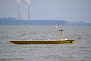 A yellow kayak-like uncrewed surface vehicle moves along Lake Erie's calm, light-greenish-brown surface. The vehicle has three dark-blue solar panels and antennae at either end. The front of the vehicle has the word SeaTrac and a right-facing dart shape printed in black. The shoreline, buildings, two smokestacks, and blue sky are in the background, out of focus in the distance on the horizon.