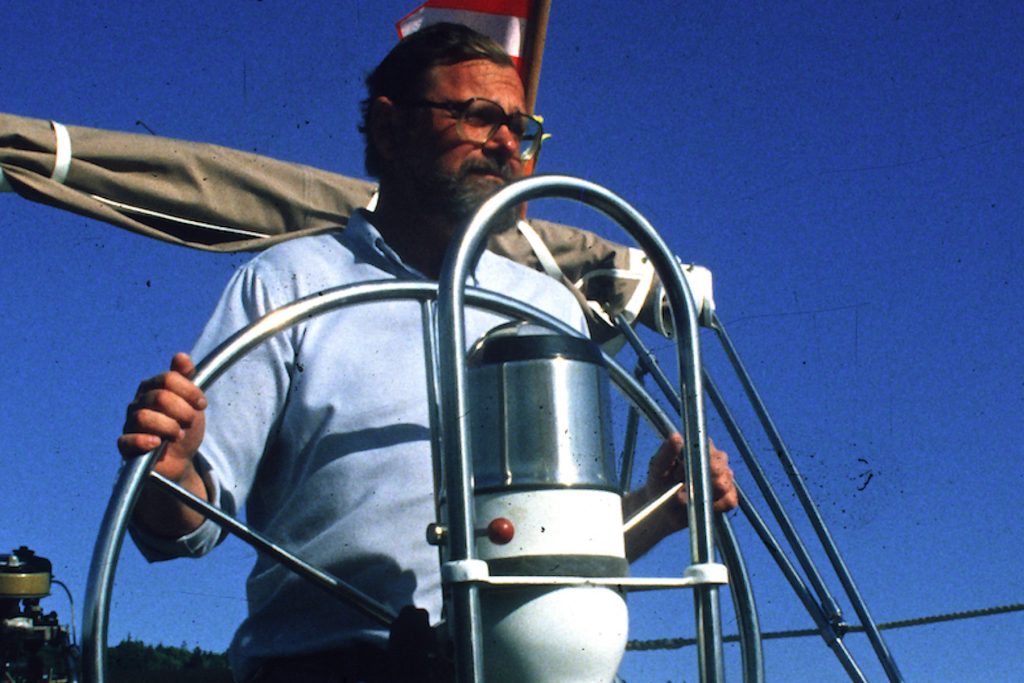 A marine biologist with dark-gray hair and a dark-gray beard wearing glasses, a light-blue shirt, and blue jeans pilots a sailboat. The biologist is holding the sailboat’s silver metal helm with two hands. Behind the biologist are two white-and-red sails flapping in the wind and a rolled-up gray sail. The background is clear blue sky.