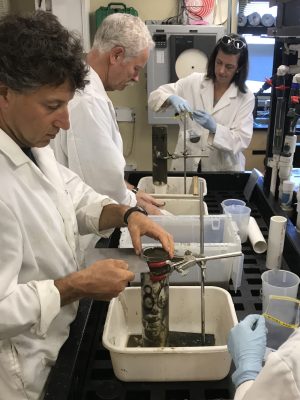 Three MBARI researchers wearing white lab coats examine specimens of brown mud in the lab aboard an MBARI research ship. The researcher on the left has short curly hair. The researcher in the center has white hair and a white mustache. The researcher on the right has shoulder-length brown hair, is wearing light-blue latex gloves, and has sunglasses resting on her head. The two cylindrical mud samples are suspended from silver metal rods placed inside rectangular, white, plastic tubs. There is a variety of scientific equipment in the background.