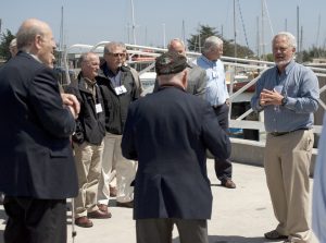 Five members of MBARI’s Board of Directors speak with two MBARI staff members on a dock. Two Board members in the center of the frame are wearing dark jackets and khaki pants with brown shoes. Two Board members in the foreground are wearing navy blazers and dark gray pants. The MBARI staff member in the center is wearing a light blue shirt, navy blue pants, and brown shoes. The MBARI staff member on the right is wearing a gray-blue shirt, khaki pants, and brown shoes. In the background are several ships docked in Moss Landing harbor, with a light blue sky overhead.