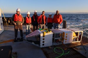 Six MBARI researchers stand on the deck of a research vessel behind a large scientific instrument. Three researchers are wearing orange life vests, two are wearing orange jackets, and four are wearing white hard hats. The scientific instrument has a rectangular silver metal frame, white plastic panels, a silver metal sphere, and a yellow plastic sphere and is sitting on the gray metal deck of the research vessel. In the background is blue ocean, with coastal mountains and blue sky in the distance on the horizon.