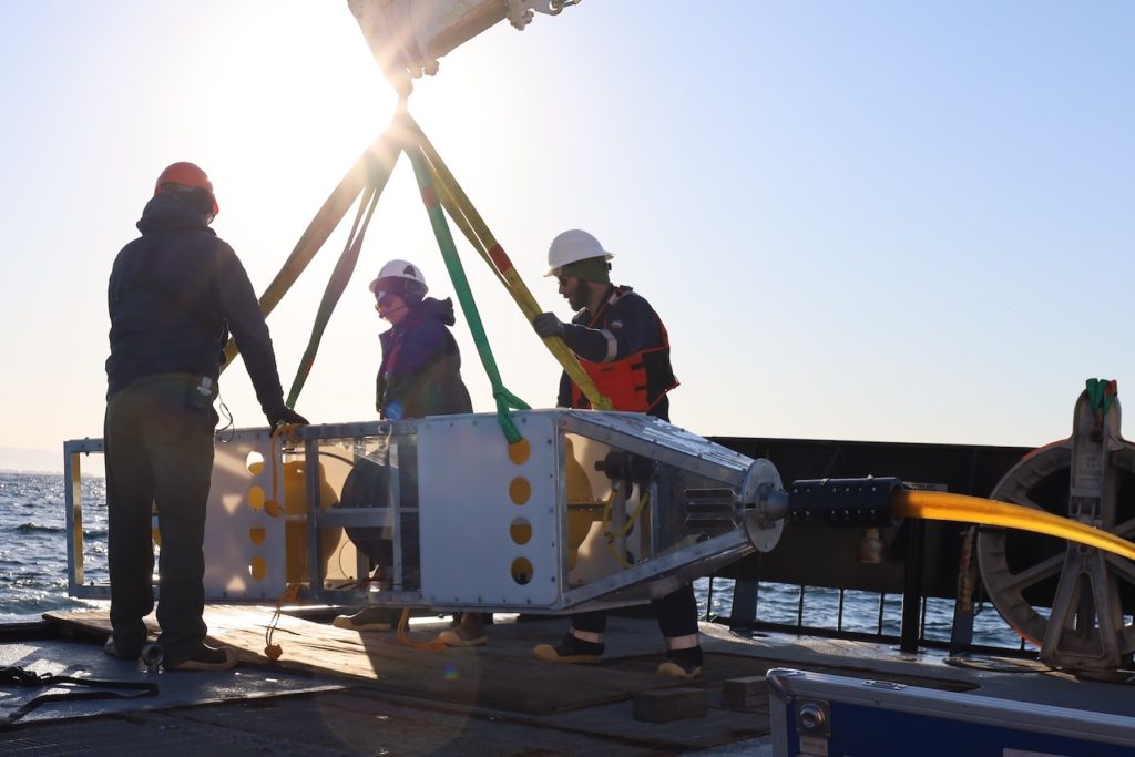 Three MBARI staff prepare for deployment of a scientific instrument on the gray metal deck of a research vessel. The researcher on the left is wearing an orange hard hat, a black hooded sweatshirt, and dark pants. The deckhand in the middle is wearing a white hard hat, sunglasses, and a dark hooded jacket. The engineer on the right is wearing a white hard hat, sunglasses, a navy-blue jacket, an orange life vest, navy-blue pants, and brown boots. All three are in silhouette in front of the bright sun. The scientific instrument has a rectangular silver metal frame, white plastic panels, a silver metal sphere, and a yellow plastic sphere and is being moved with three yellow nylon straps and one green nylon strap attached to a crane out of frame. In the background is blue ocean, with coastal mountains and blue sky in the distance on the horizon.