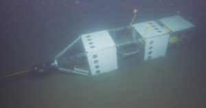 A scientific instrument with a rectangular silver metal frame, white plastic panels, a silver metal sphere, and a yellow plastic sphere rests on the muddy seafloor. A translucent orange cable connected to the instrument extends off-screen on the left side of the frame. The background is greenish-brown mud.