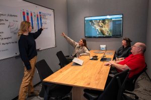 Four researchers gather around a wooden table to discuss ocean data. Seated in the foreground on the right is a researcher wearing glasses, a red polo shirt, and black pants. Seated in the background on the right is a researcher with shoulder-length brown hair wearing a navy-blue sweater. Standing in the foreground on the left is a researcher with blonde hair in a ponytail, wearing a black sweater and khaki pants, and pointing to a whiteboard. Seated in the background on the left is a researcher with long brown hair wearing an oatmeal-colored cardigan and pointing to a whiteboard. The whiteboard on the left has an illustration of data and notes from a meeting in dry-erase marker ink. The background is a gray wall with a television screen displaying a satellite map of California annotated with data points.