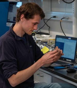 An engineer with short brown hair wearing a navy-blue quarter-zip pullover with sleeves rolled up and light-blue jeans sits at a workbench while holding up a component of research equipment. A laptop computer displaying text data is open on the black workbench counter. In the background are various pieces of engineering equipment and a white wall with black cables plugged into electrical outlets.