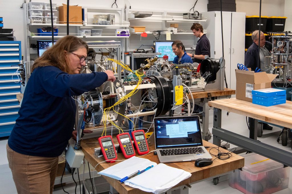 Four MBARI researchers adjust scientific instruments in a brightly lit engineering lab with white walls and shelves full of plastic bins. The researcher in the foreground has shoulder-length brown hair and is wearing glasses, a blue pullover, and khaki pants. She is adjusting a cylindrical instrument with metal components and multi-colored wires sitting next to a computer and three red voltage meters on a wooden table. The three researchers in the background are wearing blue pullovers and examining instruments with metal components and multi-colored wires.