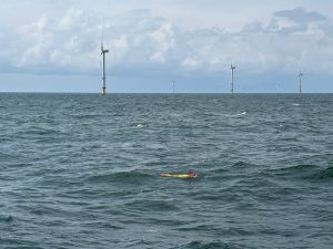 A yellow tubular underwater robot with an orange nose cone on the left and orange tail on the right moves along the surface of the ocean. In the foreground is grayish-blue ocean with choppy waves. In the background is gray sky with large white clouds. Several large white metal wind turbines are in the distance on the horizon.