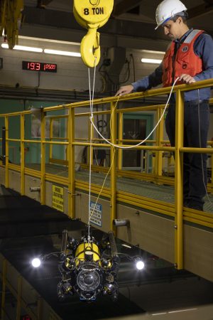 An engineer lowers a robot into a large tank of water for testing. The engineer is wearing a blue shirt, black pants, orange life vest, and white hard hat. He is standing on a beige fiberglass bridge with a yellow railing. The robot has a black metal frame and yellow cylinder metal housing with a glass-domed camera housing and two bright lights. It is dangling from white ropes attached to a large metal crane hook at the top of the frame. The background is an engineering testing lab, with the still black water of a large test tank in the foreground at the bottom of the frame.