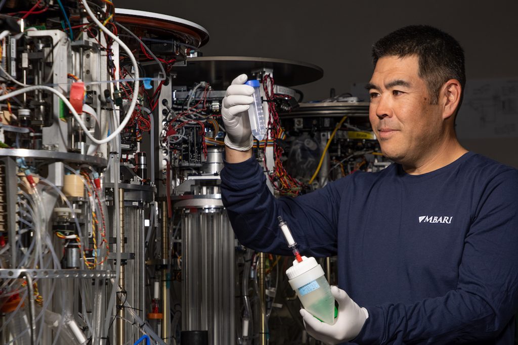 An MBARI researcher with short black hair wearing a navy-blue long-sleeved shirt with the white MBARI logo and white rubber gloves examines a clear plastic tube of fluid with his right hand while holding a white tube of fluid with his left hand. To the left are four robotic samplers with a mix of silver metal parts, multi-colored wires, and other instrumentation.