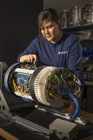 An MBARI engineer with long black hair wearing a navy-blue long-sleeved t-shirt with the white MBARI logo examines the complex robotics of a third-generation Environmental Sample Processor (3G ESP) instrument on a workbench in a research laboratory. The instrument features wiring, circuit boards, and other components around a circular metal frame. The background includes shelves holding additional equipment and robotic parts.