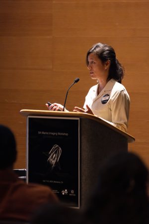 An MBARI engineer speaks into a thin black microphone at a podium. The engineer has shoulder-length black hair. She is wearing a cream-colored long-sleeved blouse and a blue-and-white name tag and is holding a laser pointer. The gray podium has a black-and-white sign with the words 5th Marine Imaging Workshop and a photo of a deep-sea comb jelly. The heads of seated conference attendees are out of focus in the foreground. In the background is a wood-paneled wall.