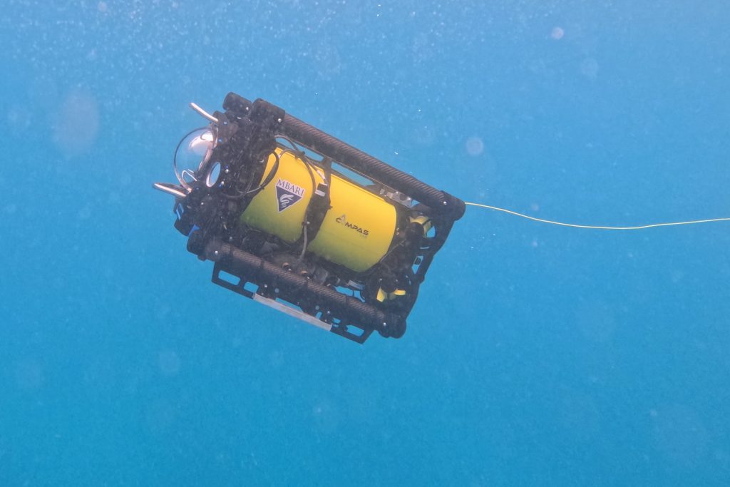 A robot moves through open water. The robot has a black metal frame outside with a clear glass dome at the front, facing the left side of the frame. Inside is a yellow metal cylinder with stickers for MBARI and the CoMPAS Lab. A thin yellow cable extends from the robot off frame to the right. The background is bright-blue water.