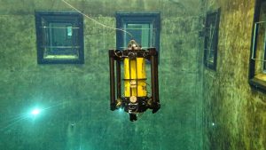 An underwater robot drifts in a large tank of water. The robot has a yellow metal cylinder surrounded by a black metal frame. The front of the robot has a glass dome camera housing and is pointed downward. In the background are gray concrete walls covered in a film of greenish-brown algae, four windows with black frames and silver metal guardrails, and two underwater lights.