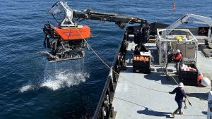 Members of MBARI’s marine operations crew recover a remotely operated vehicle after a successful dive in Monterey Bay. The robotic submersible has a bright orange float with a stylized treatment of the name Ventana that uses MBARI’s gulper eel logo as the V. Beneath the float is a black metal frame with various pieces of equipment and wires. The submersible is attached to a black-and-white crane and positioned over the side of a research ship. A bright-yellow cable connected to the submersible extends to the right to a crew member in a white hard hat, navy jacket, orange life vest, jeans, and brown boots standing on the gray deck of an MBARI research ship. A second crew member wearing a white hard hat, an orange jacket, olive-green pants, and black work boots operated a crane to the right. The background is dark-blue ocean.