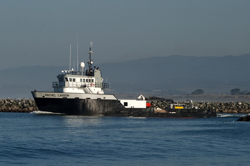 An MBARI research vessel departs the harbor. The ship has a black-and-gray hull with the name Rachel Carson in capital letters. The upper deck is white and the back deck has a white metal shipping container, orange deep-sea robot, and a large spool of yellow cable. The foreground is still blue ocean water. The background is a jetty with brown rocks, with two green trees, dark hills, and hazy sky in the distance.