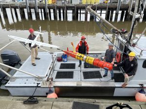 Four MBARI engineers pose for a photo on the silver metal deck of a research vessel while preparing for the deployment of advanced robots for studying the ocean. On the left is an engineer wearing a red hard hat, gray jacket, beige pants, and gray shoes and holding a white plane-shaped drone. In the center a crane holds a suspended red-and-orange torpedo-shaped underwater robot. Standing around the robot are an engineer wearing a red hard hat, a black-and-orange jacket, blue jeans, and black shoes, an engineer wearing a gray hard hat, gray jacket, blue jeans, and black boots, and an engineer wearing a yellow hard hat, black hooded sweatshirt, beige pants, and brown boots. The brownish-green water of the harbor and pilings from the gray dock are visible in the background.