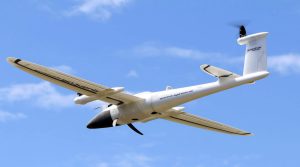 A white aerial drone flies through the sky. The drone has a black nose cone and a white body with two large wings and three black propellers. The background is blue sky with scattered white clouds.