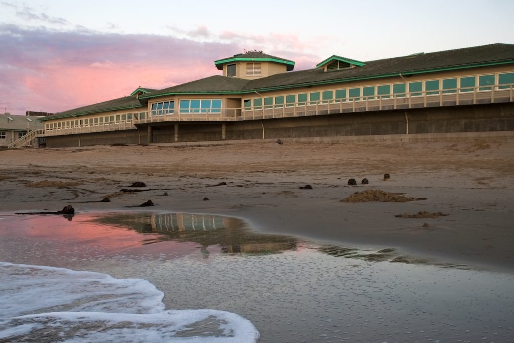 Ocean waves gently crash in front of MBARI’s research facilities. The research facilities have gray concrete walls, several large windows, and a gray roof with teal trim. In the foreground are wet sand and several clumps of brown drift kelp. In the background is a pale blue sky with pink clouds.