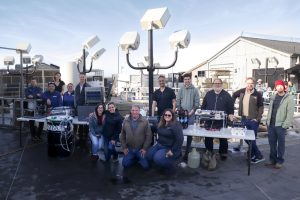 Fourteen researchers pose around five eDNA sampling instruments placed on gray tables during an experiment at the Monterey Bay Aquarium. Behind the researchers are tall white spotlights on black metal poles and various aquarium life support infrastructure, with blue sky and white haze in the background.