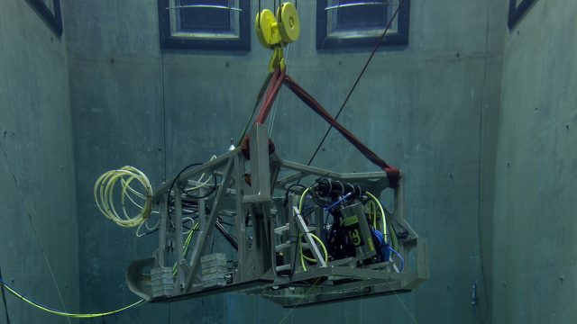 The seafloor mapping toolsled hangs from a crane inside MBARI’s test tank. The toolsled has a weathered metal frame, silver and black metal canisters, and a tangle of green, blue, and yellow cables. The toolsled is suspended by a yellow crane hook and red ropes as the lidar canister projects three green lasers toward the bottom of the tank. The background is the gray concrete wall of the test tank.