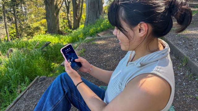 A person sitting in a park plays the FathomVerse game on their mobile phone. The person has shoulder-length black hair in a ponytail and is wearing a sleeveless sweater with beige and white stripes and blue jeans. They are holding a smartphone in a pink case. The person is outdoors and in the background on the right are terraces of brown dirt, and on the left are green grass and brown tree trunks.