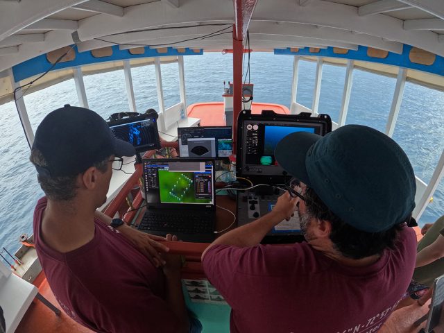 Two engineers wearing navy-blue caps, glasses, and maroon t-shirts standing in the cabin of a wooden boat look at computer screens displaying data from an underwater robot. The engineer on the right holds a controller and steers the underwater robot. Four computer screens sit in black cases on a table in front of the engineers in the center of the boat. The background is the deck of the boat with open-air windows and blue ocean.