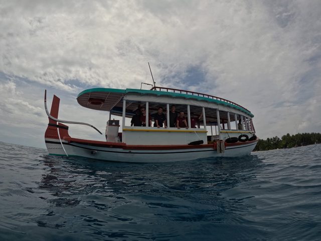 A white wooden dhoni boat with a teal-green roof and brick-red bow drifts in open water. Seven silhouetted crew members are standing aboard the boat, leaning against the railing of the cabin and looking at the camera through open-air windows. The dhoni is in the center of the frame with blue-gray water in the foreground. In the background are an island with green trees and cloudy gray sky.