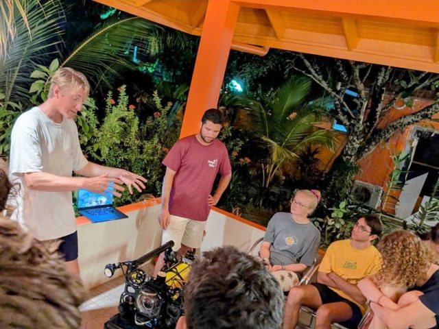 Two engineers speak with a group of students on the veranda of a marine research station. The engineer on the left is wearing a white shirt and navy shorts, and the engineer on the right is wearing a maroon shirt and khaki shorts. In front of the engineers is an underwater robot with a yellow metal cylinder, glass dome camera housing, and a white headlight, all surrounded by a black metal frame. Students are seated around the engineers. The veranda has a white half-wall, orange column, and orange ceiling, with open-air windows and green tropical vegetation in the background.