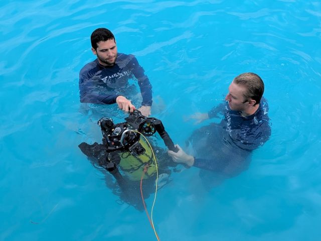 Two engineers drift at the ocean’s surface while holding an underwater robot. The engineers are wearing navy-blue rash guards with the white MBARI logo. The robot has a yellow metal cylinder surrounded by a black metal frame, black propellers, and electronics. A thin yellow cable and a thin orange cable extend out of frame to the bottom. The background is bright-blue water.