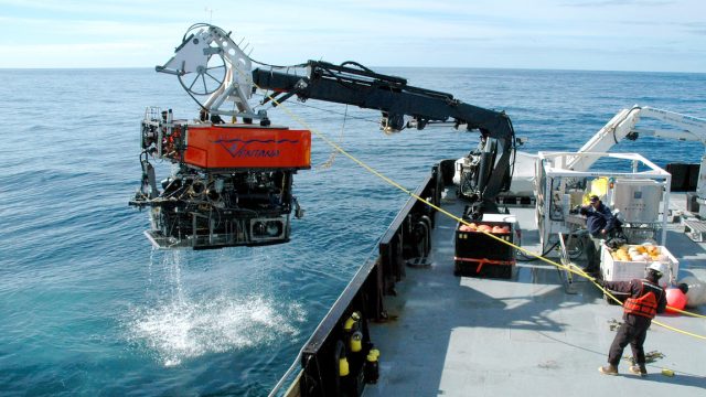 Members of MBARI’s marine operations crew recover a remotely operated vehicle after a successful dive in Monterey Bay. The robotic submersible has a bright orange float with a stylized treatment of the name Ventana that uses MBARI’s gulper eel logo as the V. Beneath the float is a black metal frame with various pieces of equipment and wires. The submersible is attached to a black-and-white crane and positioned over the side of a research ship. A bright-yellow cable connected to the submersible extends to the right to a crew member in an orange jacket, black pants, and brown boots standing on the gray deck of an MBARI research ship. The background is bright-blue ocean and blue sky.